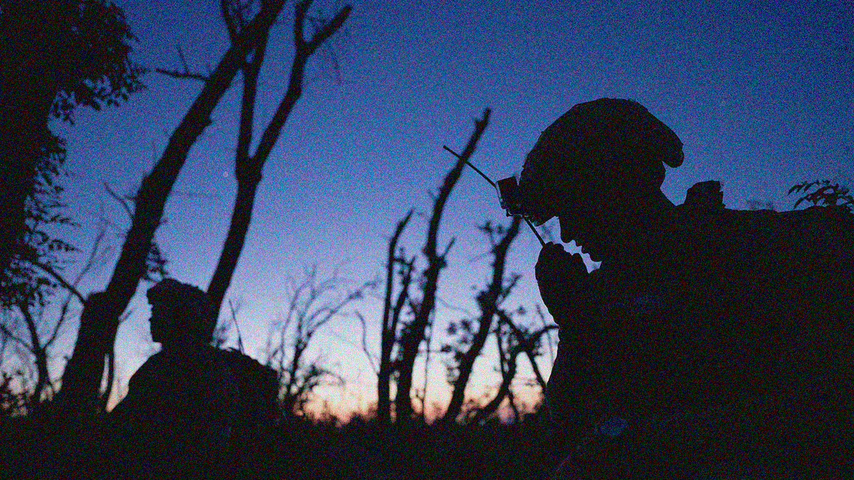 Night scene of two Ukrainian soldiers in silhouette amidst a barren forest. One holds a radio receiver to his mouth.