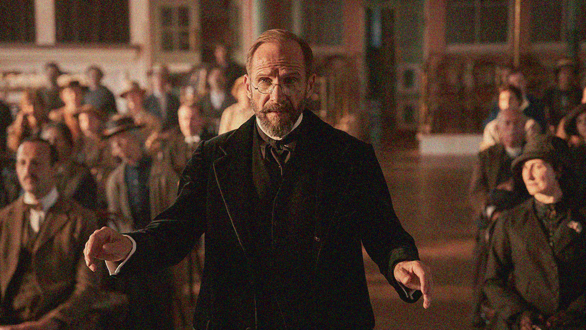 A bearded and bespeckled man in suit coat stands center frame with arms out in conducting position with an audience behind him.