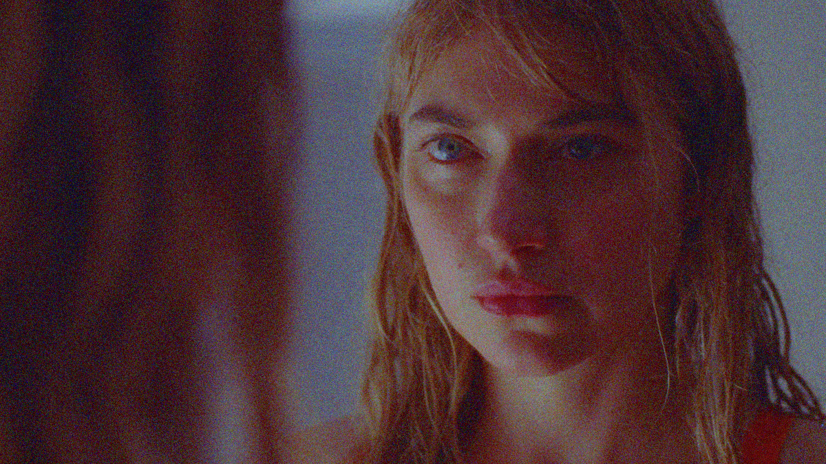 Close-up of the reflection of a blonde woman's face caught in a severe stare. We see her hair in the foreground at left to show the full face is in a mirror.