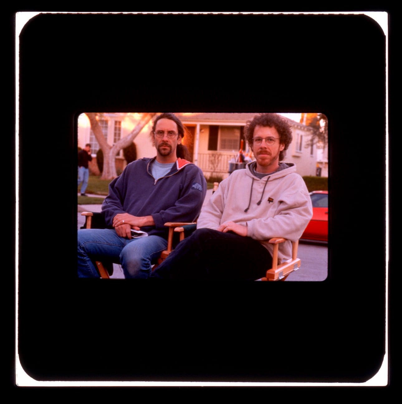 Color publicity slide: Two men in glasses sit in director chairs on a residential street. One has long hair and a goatee and wears a quarter zip hoodie. The other has full beard and curly hair and wears a full hoodie.