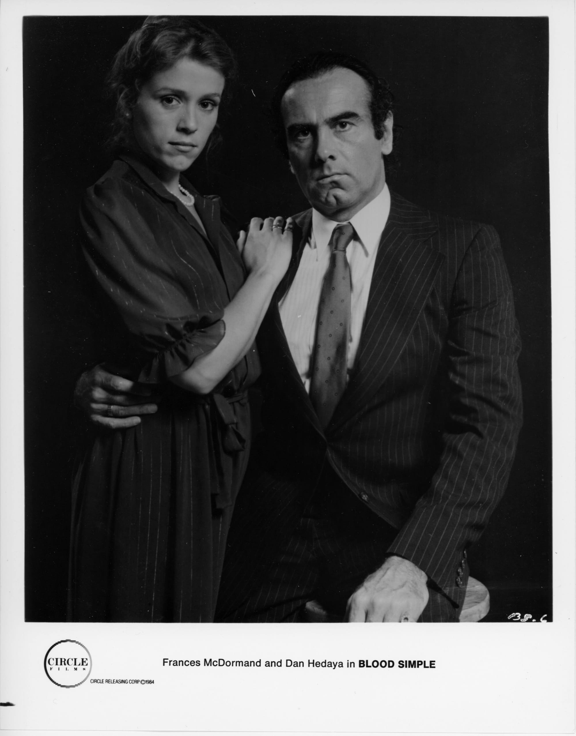 B&W publicity photo: Portrait of woman and man. He is in suit and tie sitting on a wooden stool with head cocked, hand around her waist. She stands by his side in dress and pearls, hand on his shoulder. Both look stoic, no smiles.