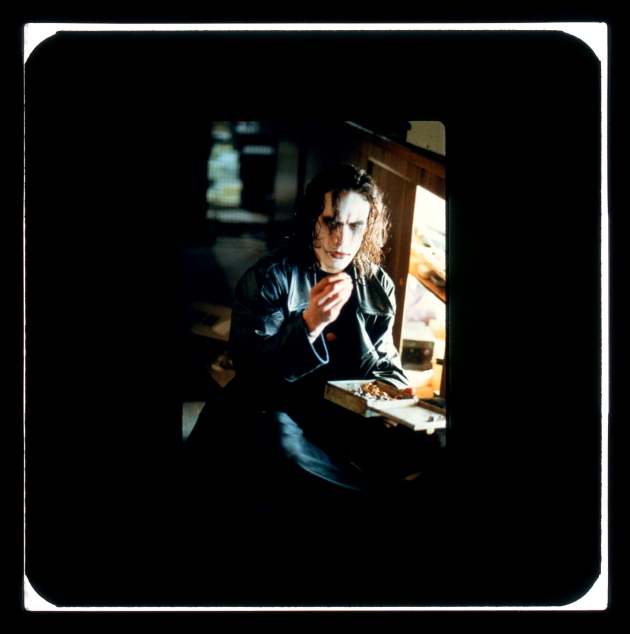 Color publicity slide featuring Brandon Lee as the Crow in a leather jacket, white face paint, red lips, and black around eyes, long hair in face. He is looking at something in his hand.