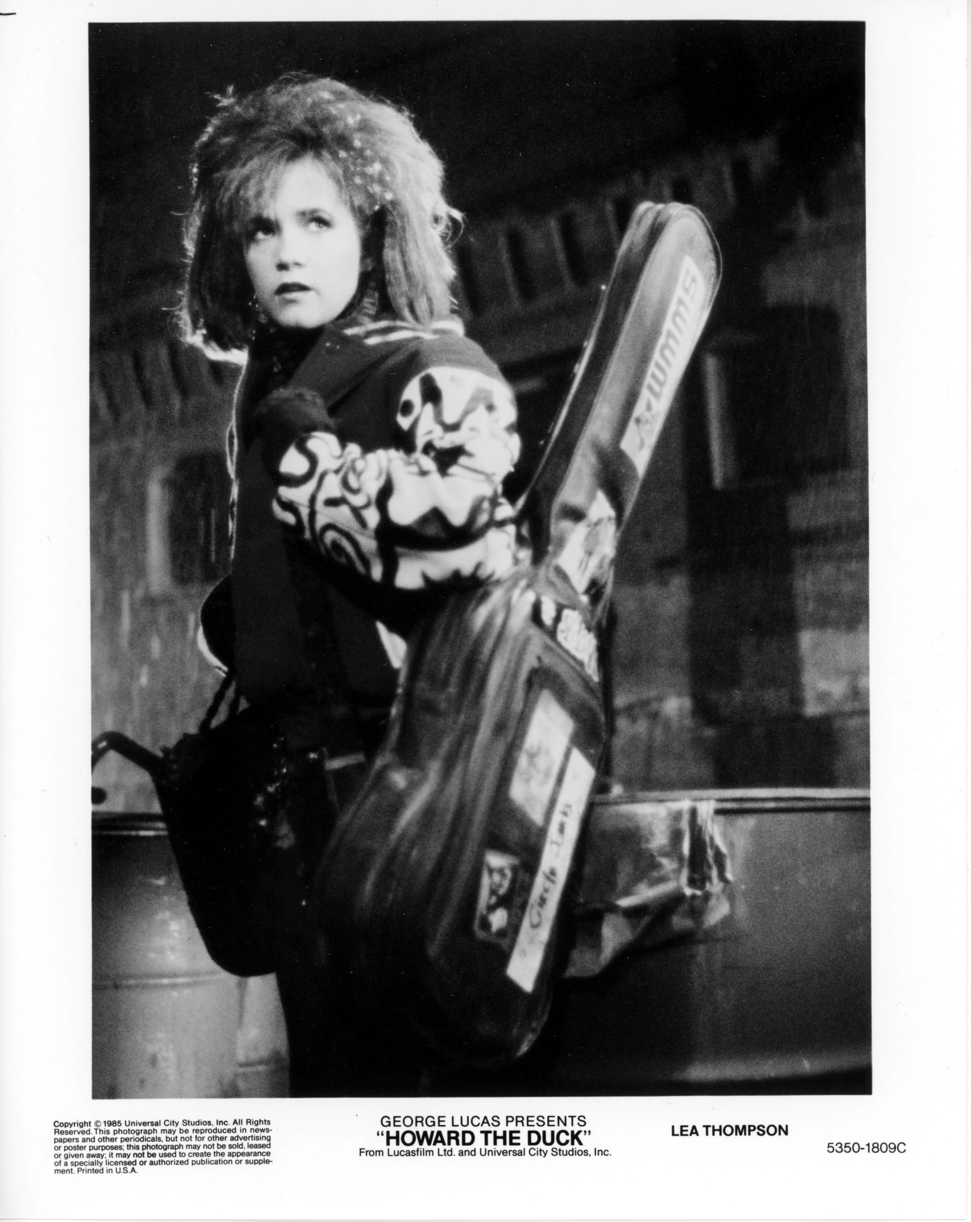 B&W Publicity Photo: A woman stands outside with guitar case slung over shoulder facing left while looking off-screen right.