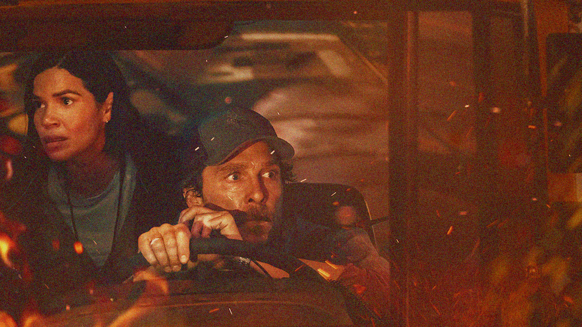 A man in ball cap is behind the wheel of a bus center frame with a woman standing behind his right shoulder and almost cropped off the left side of the frame. The whole filtered yellow with embers flying at the bottom and right half of photo.