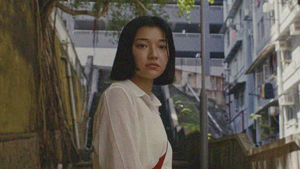 A woman in white stands center frame outside. City buildings are seem behind her.