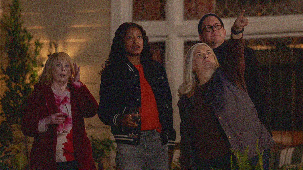 Four people stand outside the front of a home at night looking up and off-screen right with looks of shock. One points towards where the others are looking.