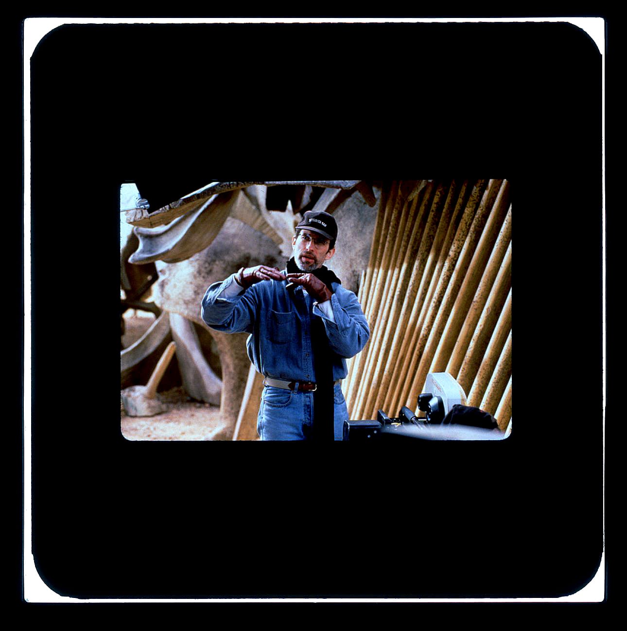 Color Publicity Slide: A man in jeans and jean button-down wears a black hat and glasses. His hand are up palms down and fingers touching as he explains something to people off-screen.