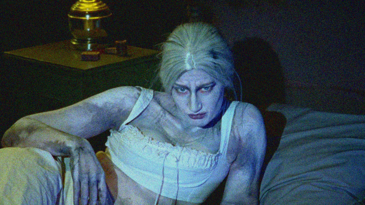 A pale woman (in white bra and covered in dirt and grime) lays on a bed with arm on hip, staring into the camera.