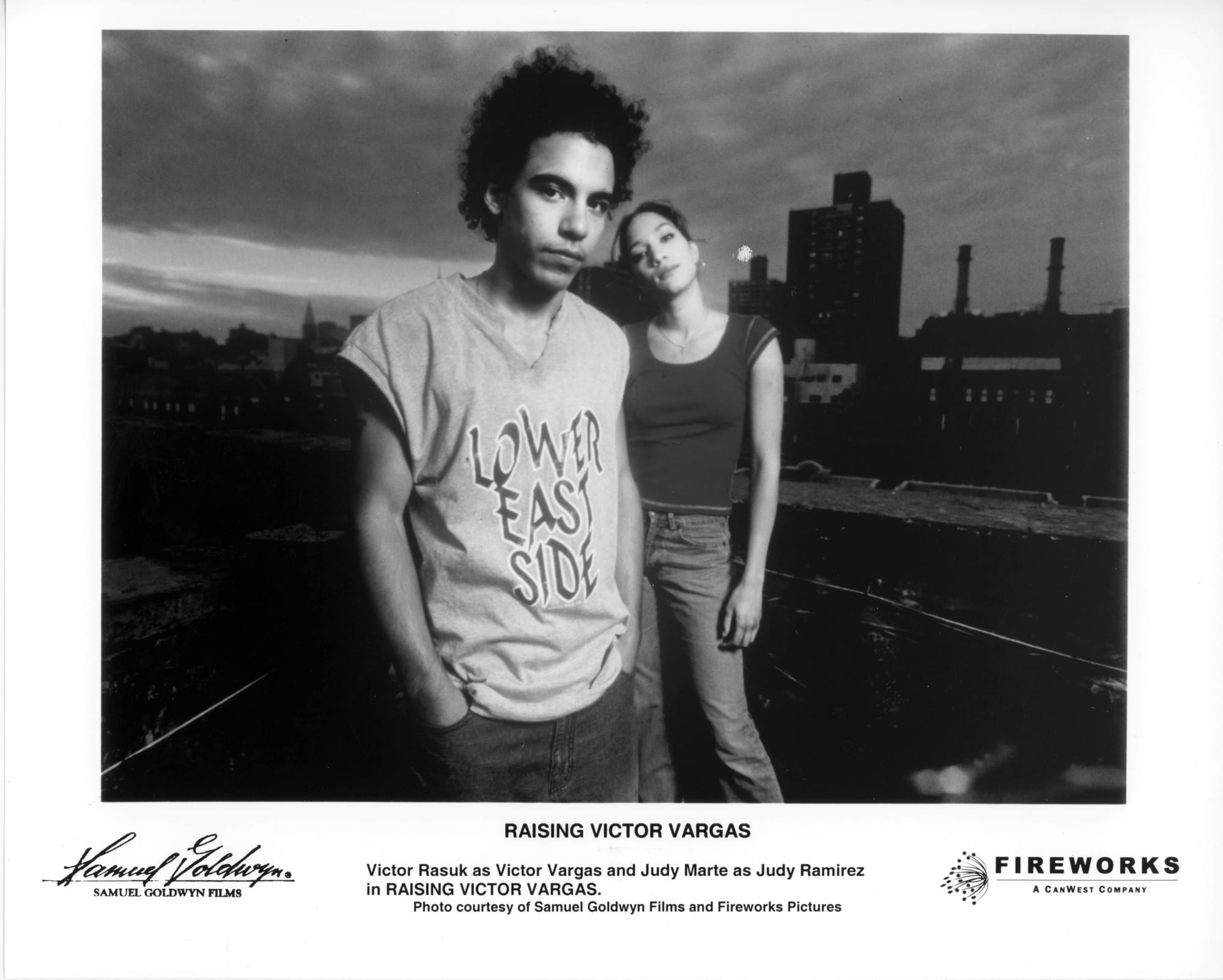 B&W Publicity Photo: A young man in a sleeveless shirt that says "Lower East Side" and hands in pants pocket stands in front of a young woman in tank top with head tilted. It's an outdoor scene with buildings in the background.
