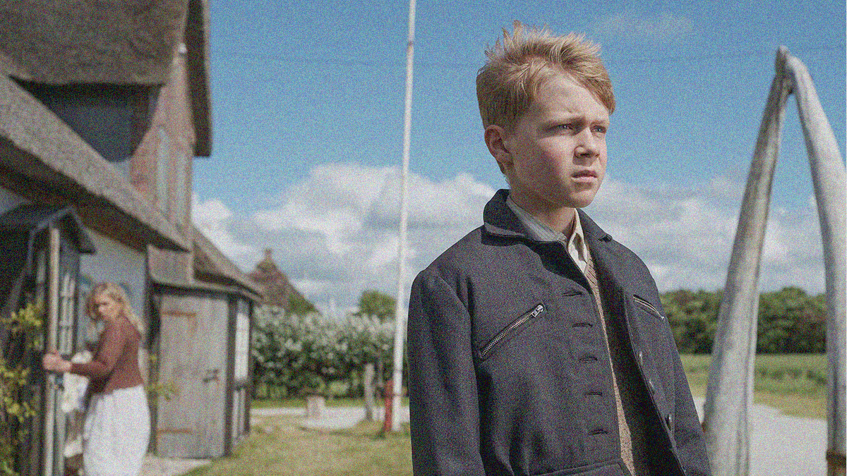 A young boy in a jacket (seen from waist up) stands in the foreground looking offscreen right with a furrowed brow. A woman is in the background look at him as she goes into a house.