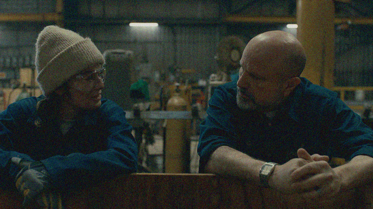 A woman on left and a man on right both lean forward onto a wood plank. Their hands are both crossed in front of them on the ledge and both are looking at each other in conversation.