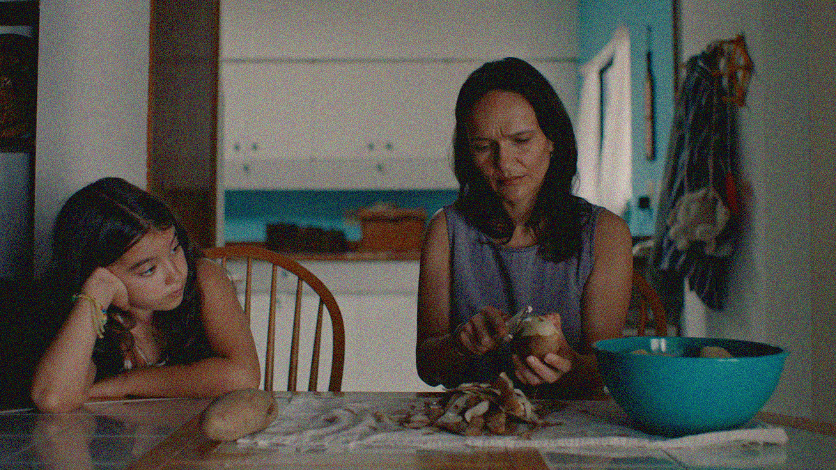 A mother and daughter sit at a kitchen table. The former is peeling a potato. The latter is watching with elbow on table and head in hand.