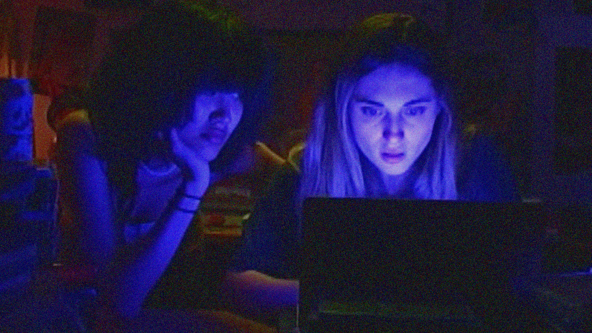 A young woman looks at her laptop screen so the blue light from it illuminates her face. Another young woman leaning her head on her hand (elbow on desk) looks at her.