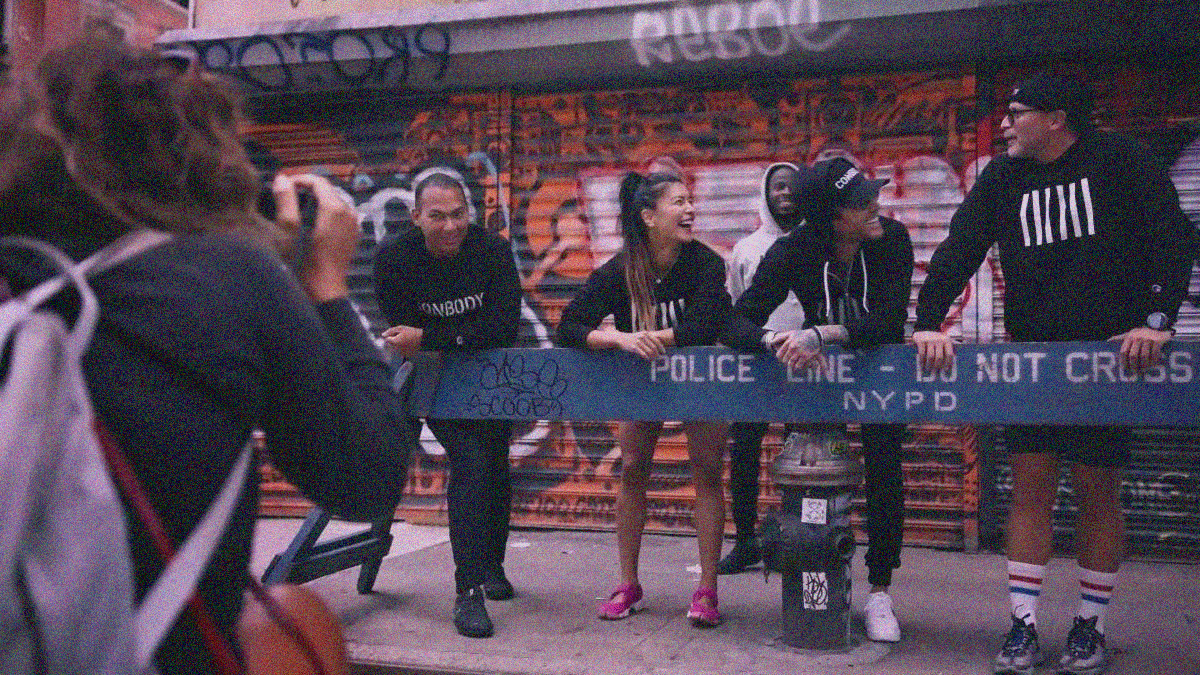 Four people in black clothing with white vertical bars or the word "Conbody" stand laughing behind a blue Police Line barrier. Graffiti store fronts are behind them. A photographer is in front of them.