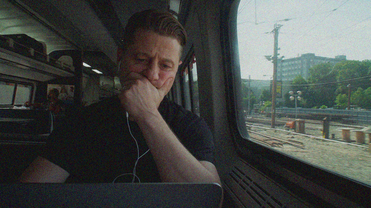 A man with headphones in ears and face smushed by hand covering mouth below nose is looking at a laptop screen. He's on a train with the window outside filling the right half of the frame.