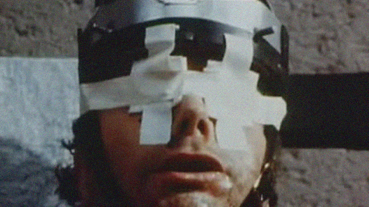 Close-up of a man with eyes covered by gauze and tape and head enclosed by an electric chair helmet.