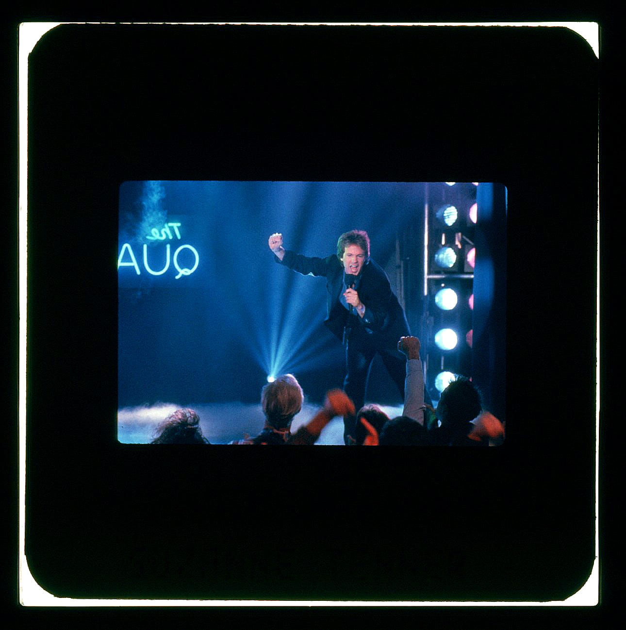 Color Publicity Slide: A man stands on a stage in blue light with one fist in the air and a microphone in the hand. He's leaning forward off the stage to sing to the crowd in front.