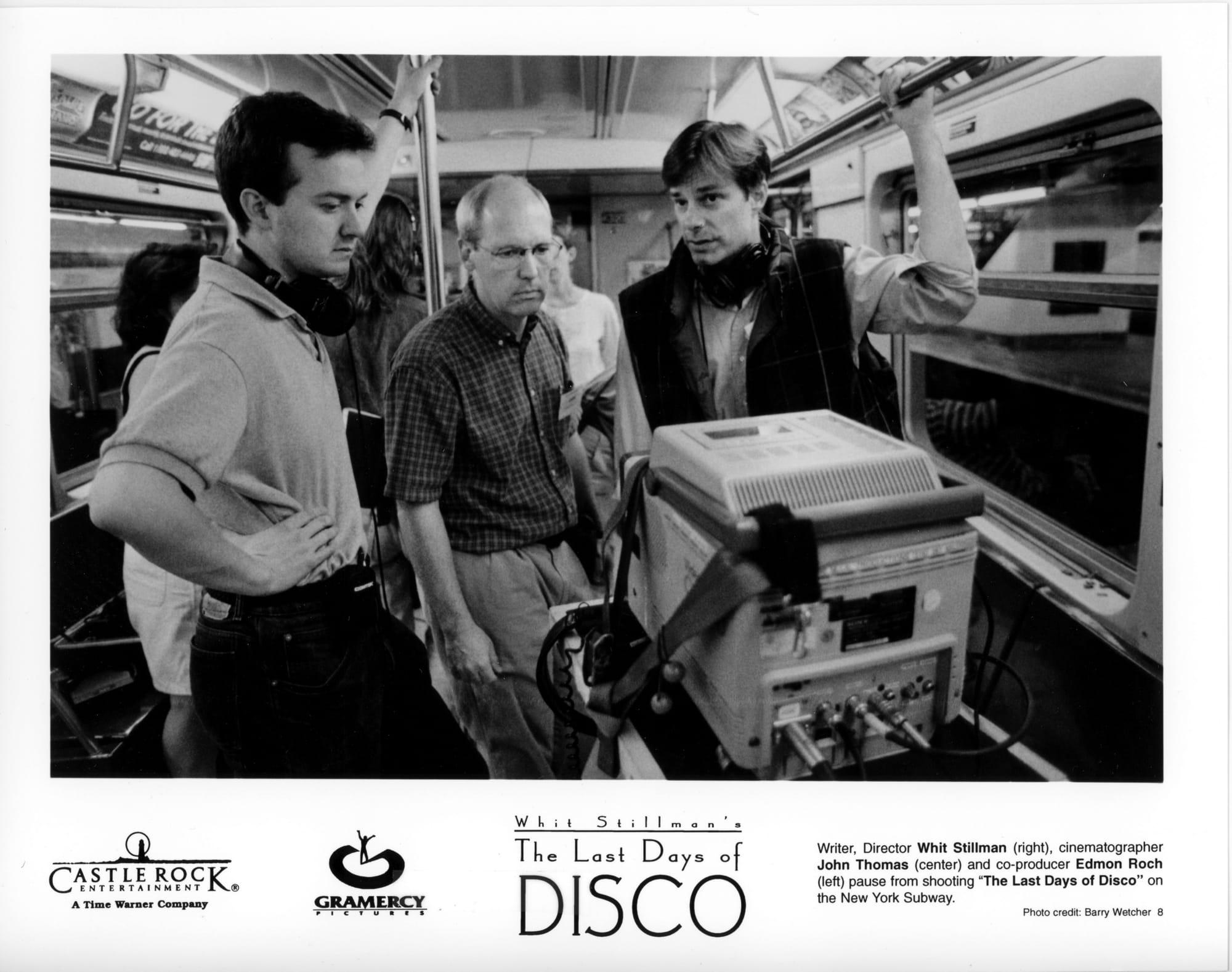 B&W Publicity Photo: Three men stand behind an electronic box presumably with a video screen while standing on set inside a subway car. The man on left and right have one hand each on metal poles. The man in the middle is looking intently at the screen.