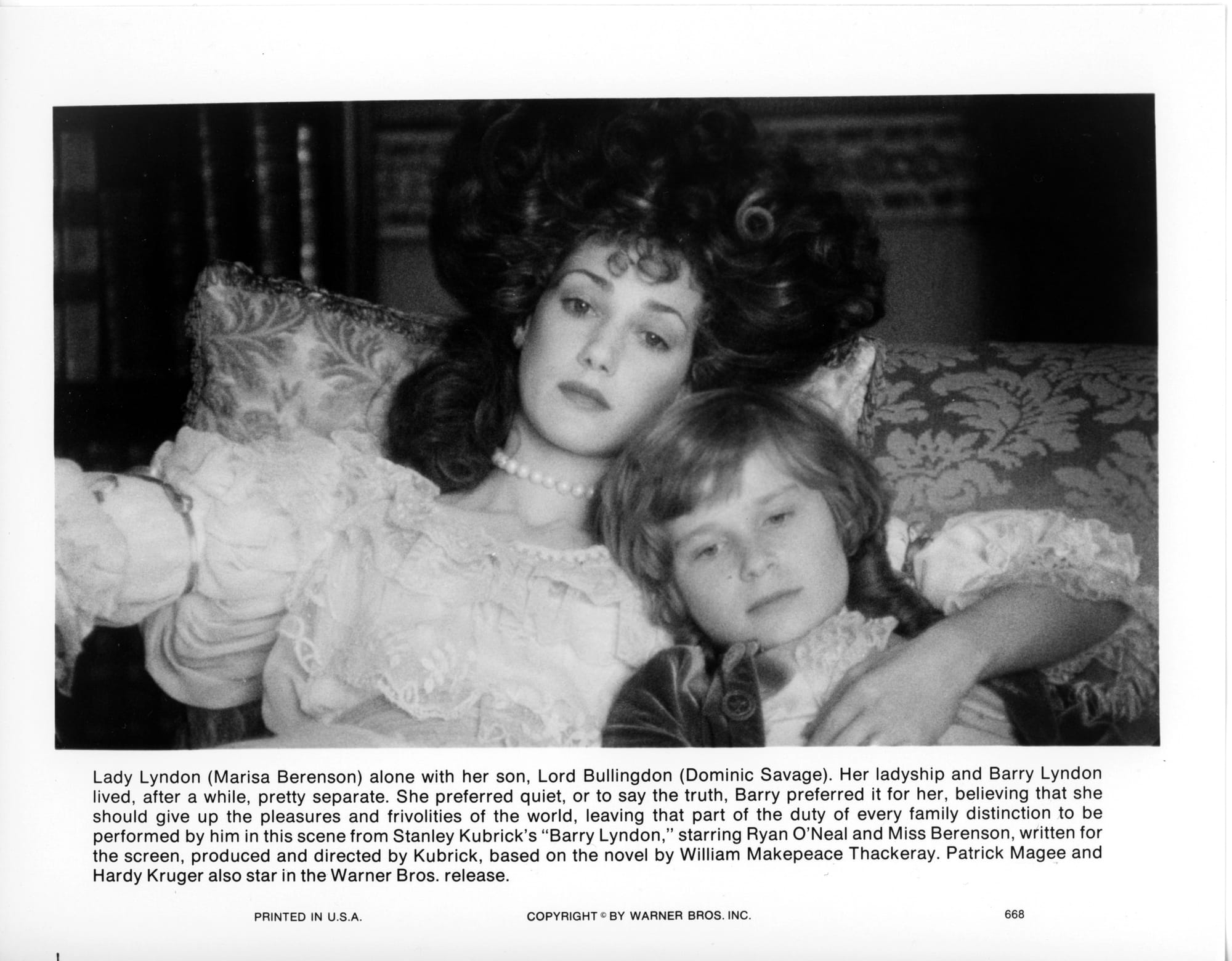 A woman with big hair in white dress lounges on a pillow with a young boy wrapped under her left arm.