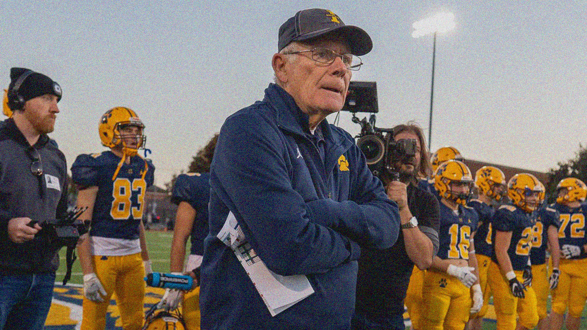 An older man with arms crossed at chest looks on as a cameraman and a team of high school football players stand behind him.