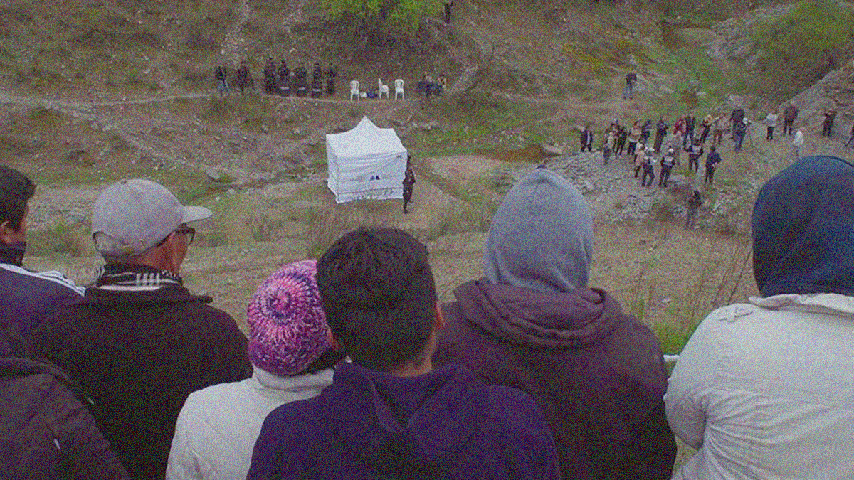 Outdoor scene on hilly terrain. A group of people are in the foreground with backs facing the camera as they watch people in the distance.