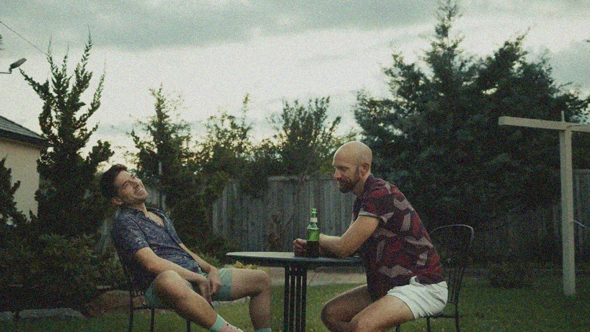 Two men sitting at a small table outside in a yard enclosed by a wooden fence and foliage. One leans back smiling. The other leans forward with bottle in-hand.