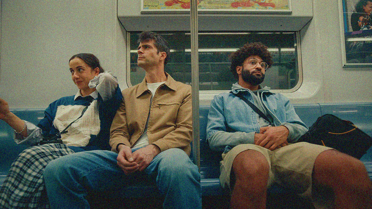 An image of a subway train bench bisected by a metal pole. A woman and man are seated on the left looking left and a man is sitting on the right looking right.