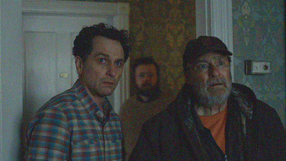 Two men stand side by side looking past the camera with scared and confused spaces. They stand in a doorway with another man and closed door in the hall behind them. 