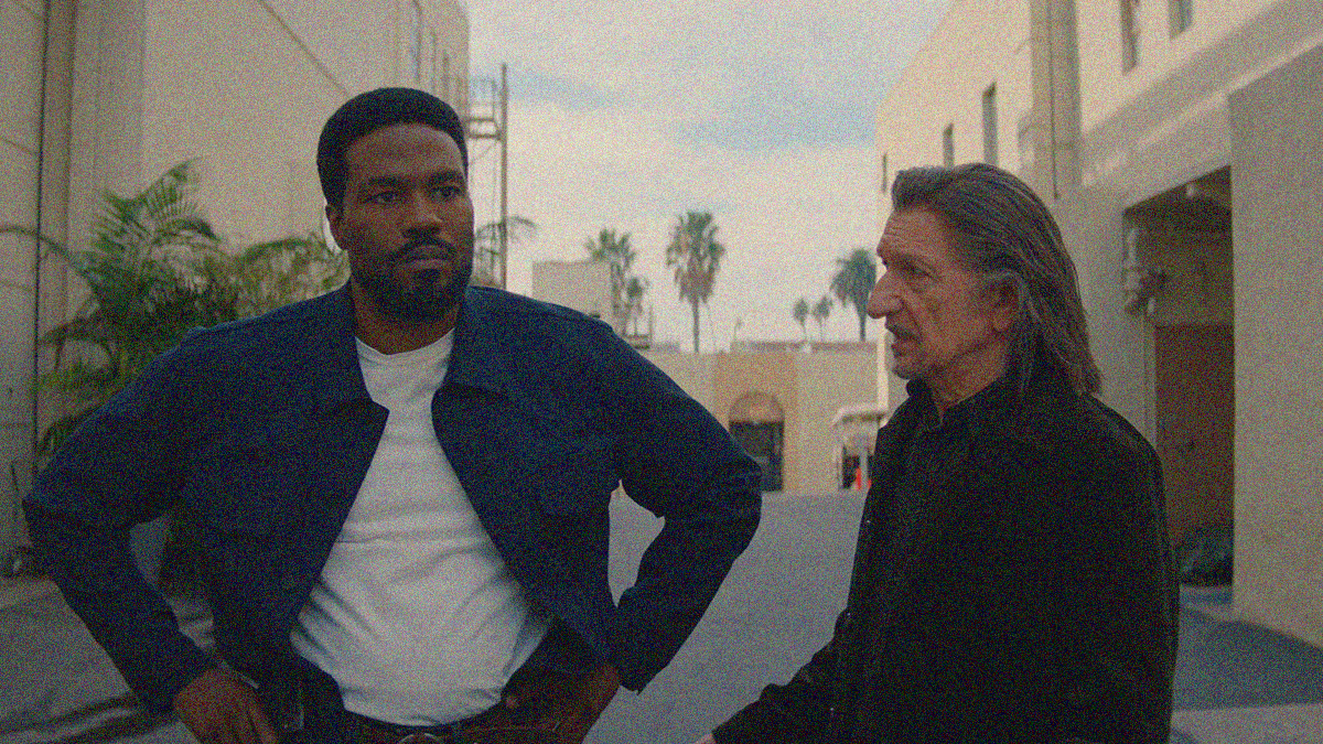 A bearded man stands with hands on hips, staring off-screen with a frown, while a long-haired man stands looking at him with mouth open in conversation.