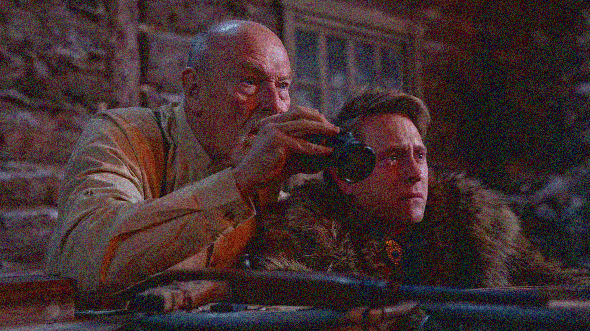 Two men stand outside a cabin and crouch by a railing. The younger one is in fur with a tear in his eye. The older one stands higher behind him with binoculars in-hand.