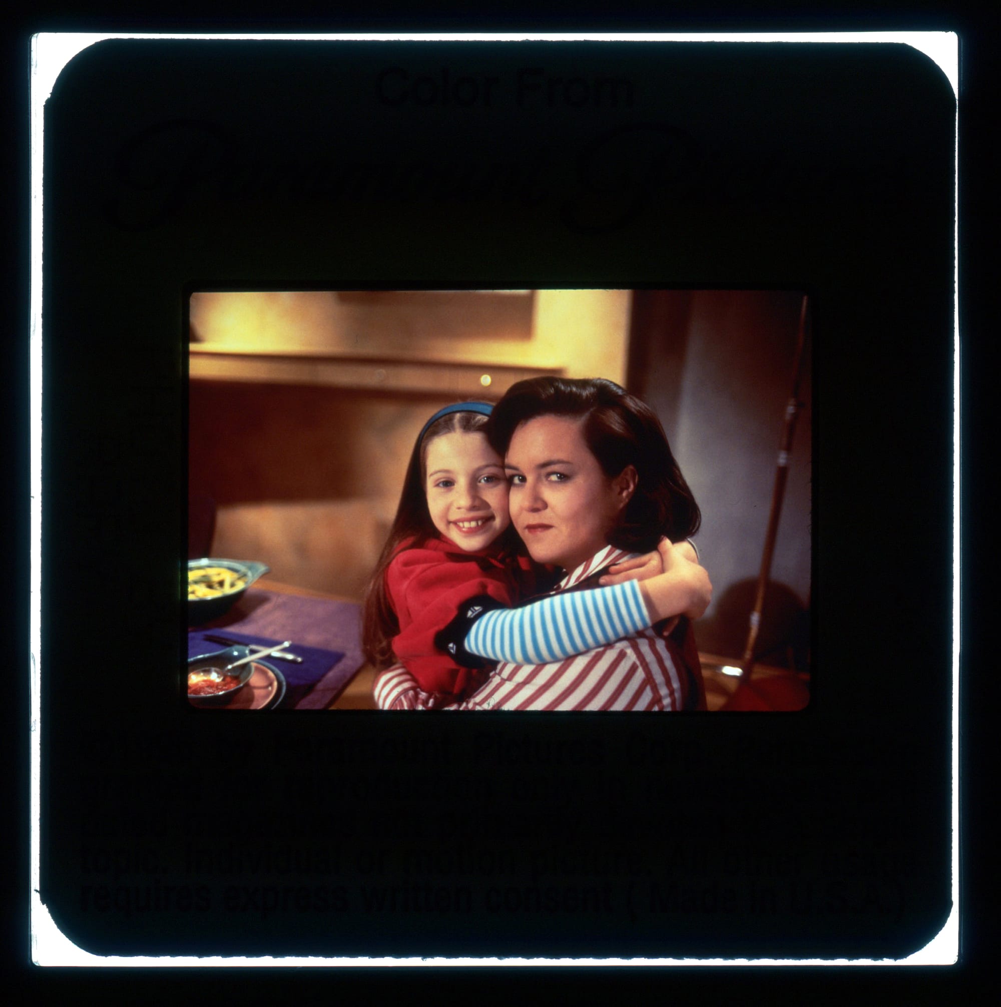 A woman sits at a table with a young girl hugging her around the neck. Both are looking and smiling at the camera.