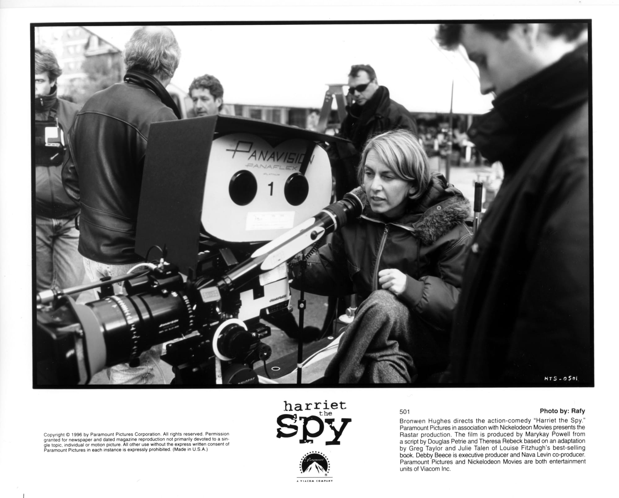 A woman sits behind a giant Panavision camera and peers down into the viewfinder while other crew members mill about.