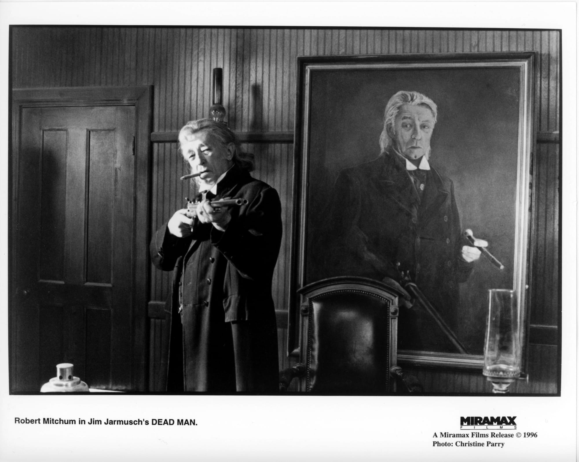 B&W Publicity Photo: Mitchum stands with cigar in mouth and shotgun pointed towards camera. A large painting is on the wall behind him of Mitchum with cigar in one hand and shotgun pointing down in the other.