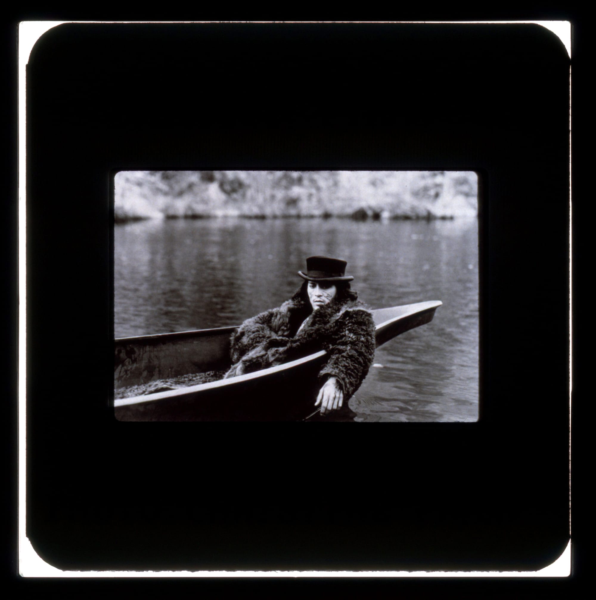 Publicity slide: Depp in a fur coat and hat with zigzags painted on his cheeks, lays in a canoe on water. One hand is under the coat, the other hangs over the water.