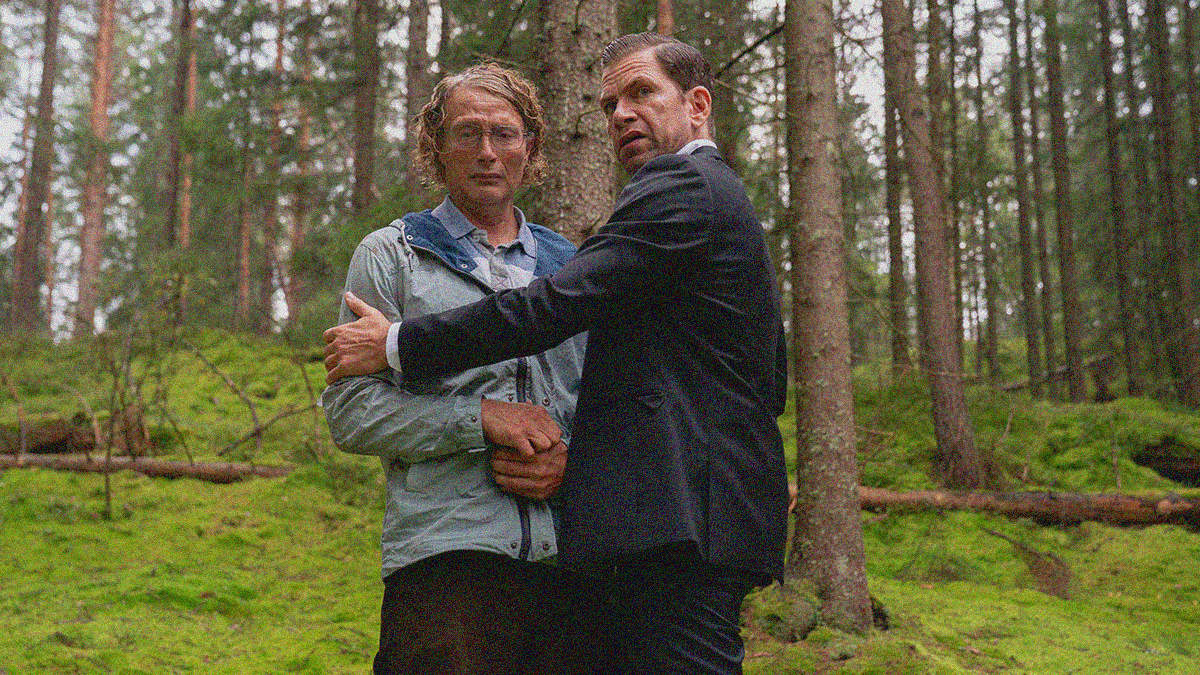 A man in a suit on right holds a sad man in a blue coat on left in a tenuous embrace. They are standing against a tree outside in the woods.