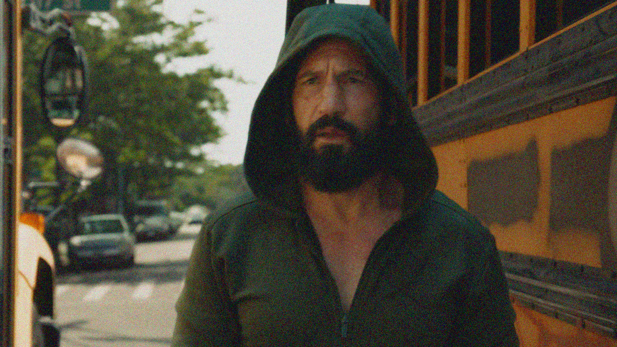 A beard man in a hoodie walks between two vandalized school buses on the street.