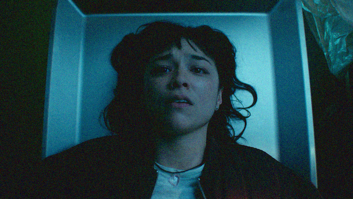Close-up of a young woman lying on a morgue slab looking up with a worried expression. The camera angle is from above to just see her from shoulders up.