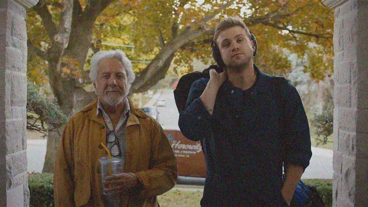 An older man (on left with a plastic drink cup in-hand) and a younger man (wearing headphones and holding a bag over his shoulder) face the camera as though we are the front door of a house.