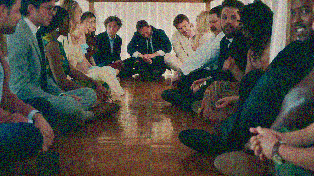 A large collection of adults in wedding attire are sitting crosslegged underneath and along the interior perimeter of a table.