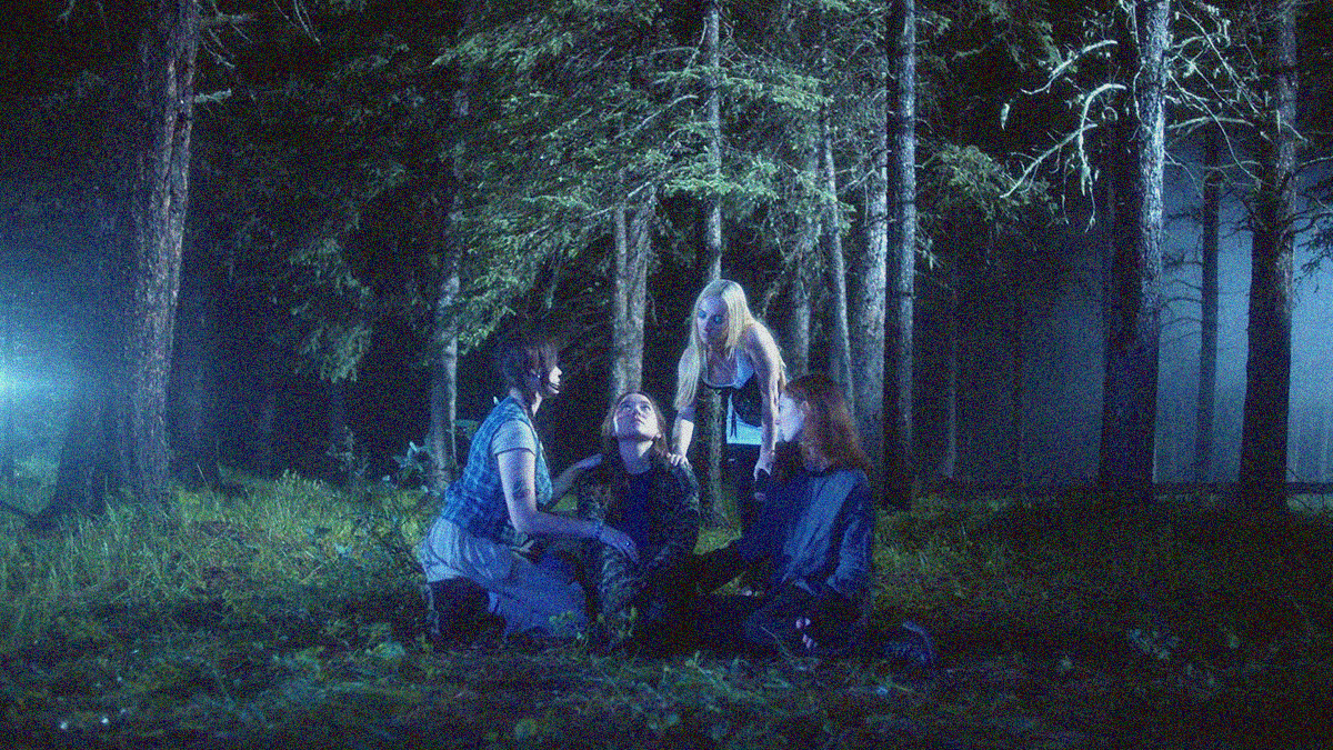Three women surround a fourth in the woods as she looks up into the sky.