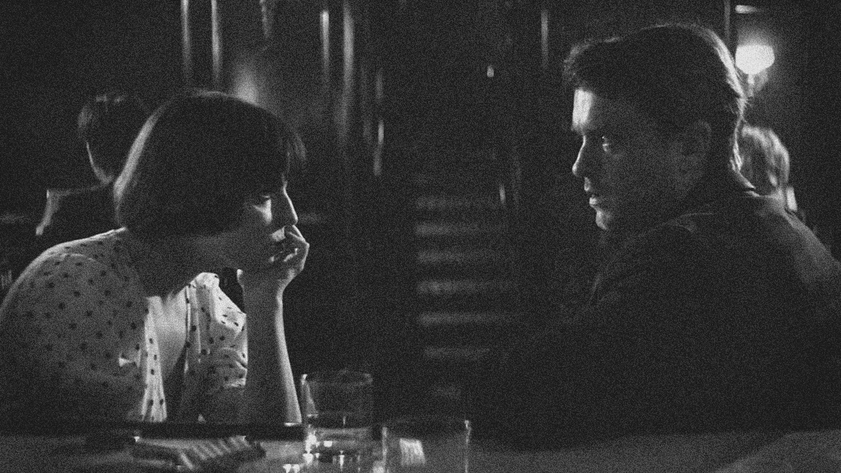 A woman with chin propped on hand and a man with arm on bar sit and face each other inside a darkened club.