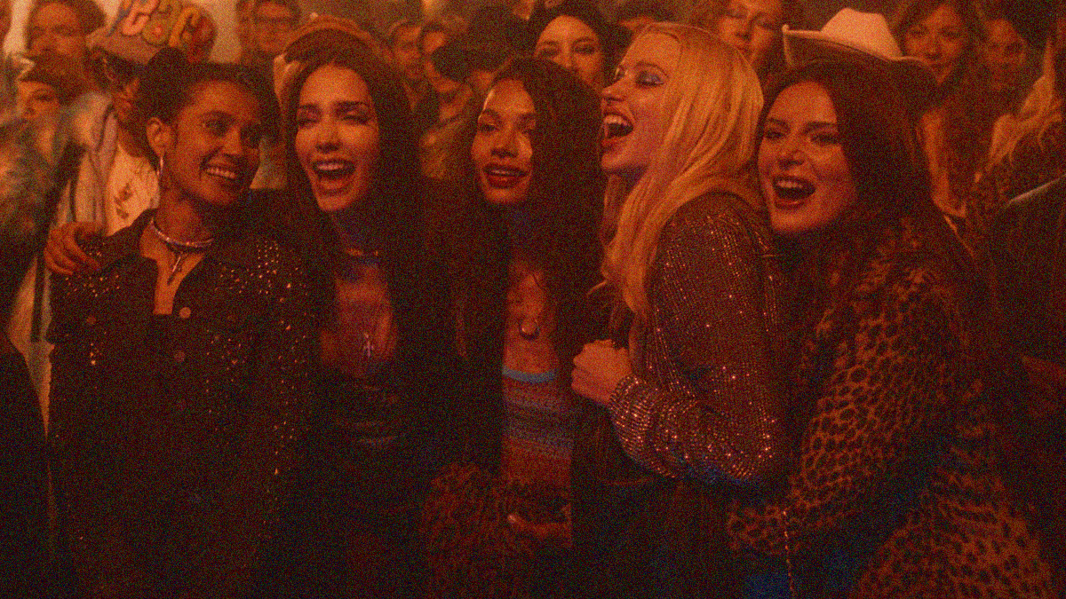 Five women has their arms around each other while screaming and smiling in a crowd of people at a concert.