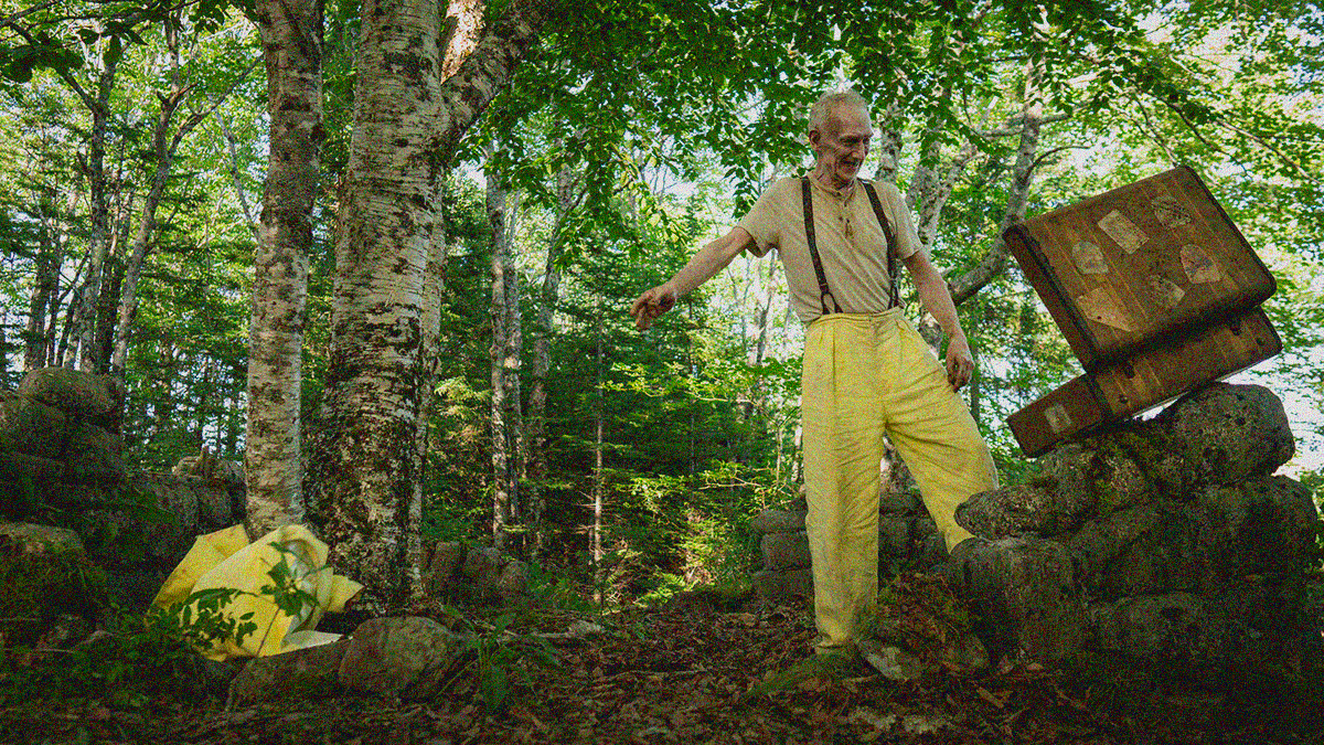 A tall man in yellow pants with suspenders looks like a battered open suitcase after throwing his yellow suit coat onto the dirt in the woods.