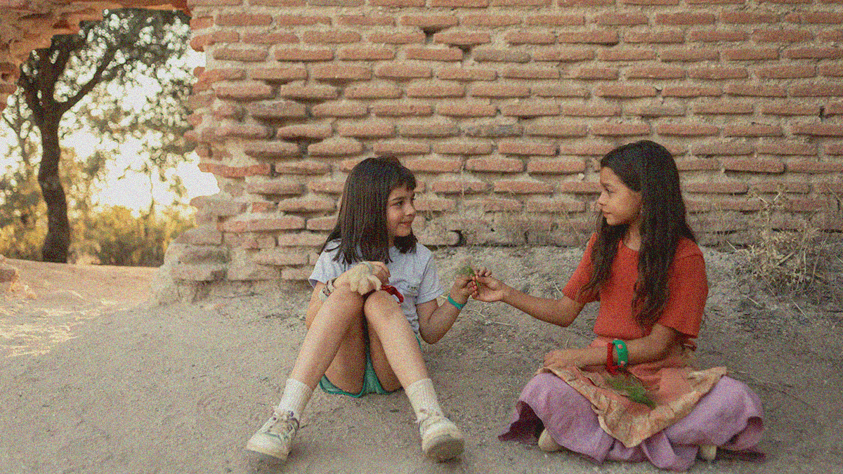 Two young girls sit on the dirt ground against a brick wall outside. Each has one outstretched hand reaching to the other's while smiling.