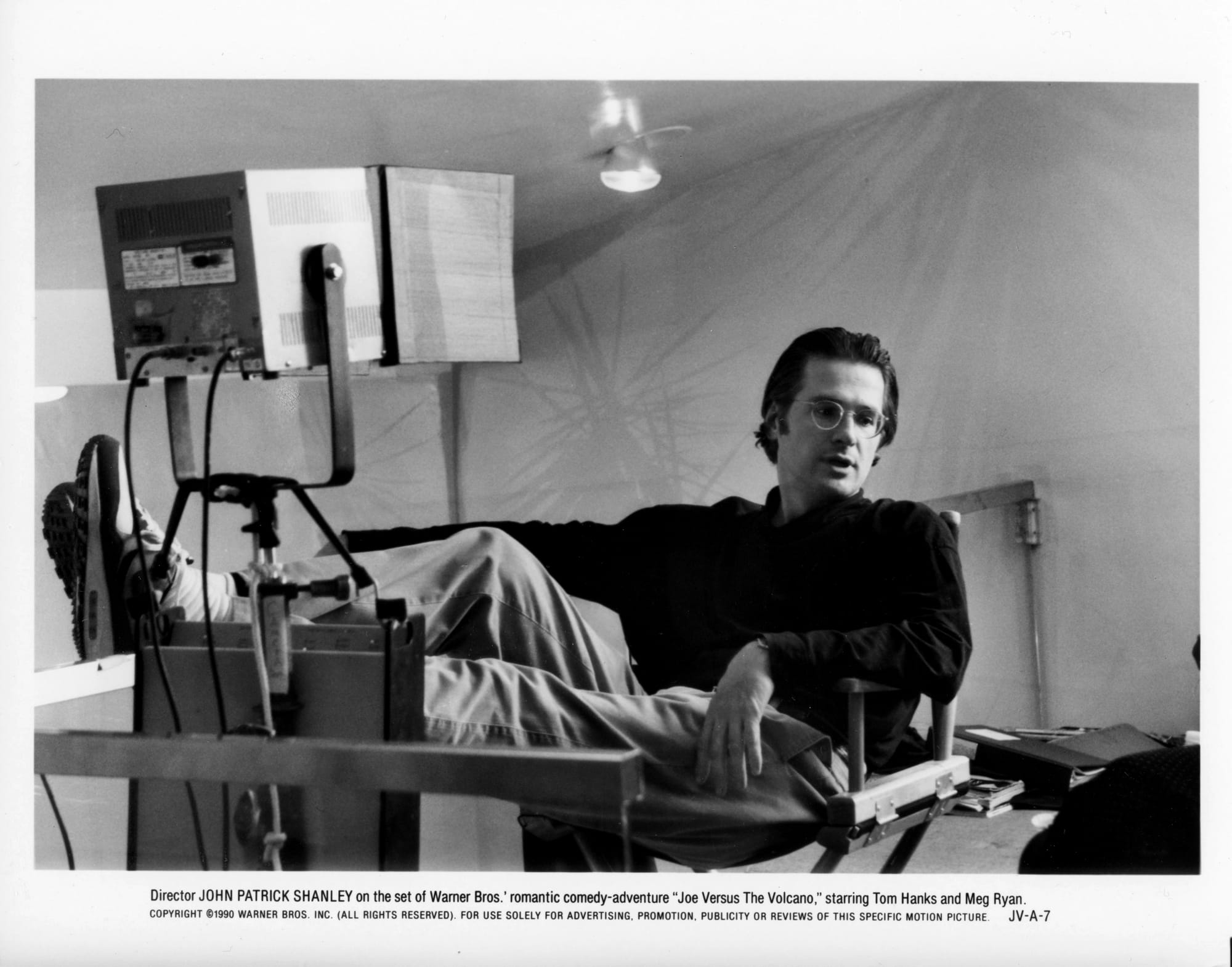 Black and White Publicity Photo: A man in glasses lounges in a director's chair with legs outstretched and up on a light rig in front of him.