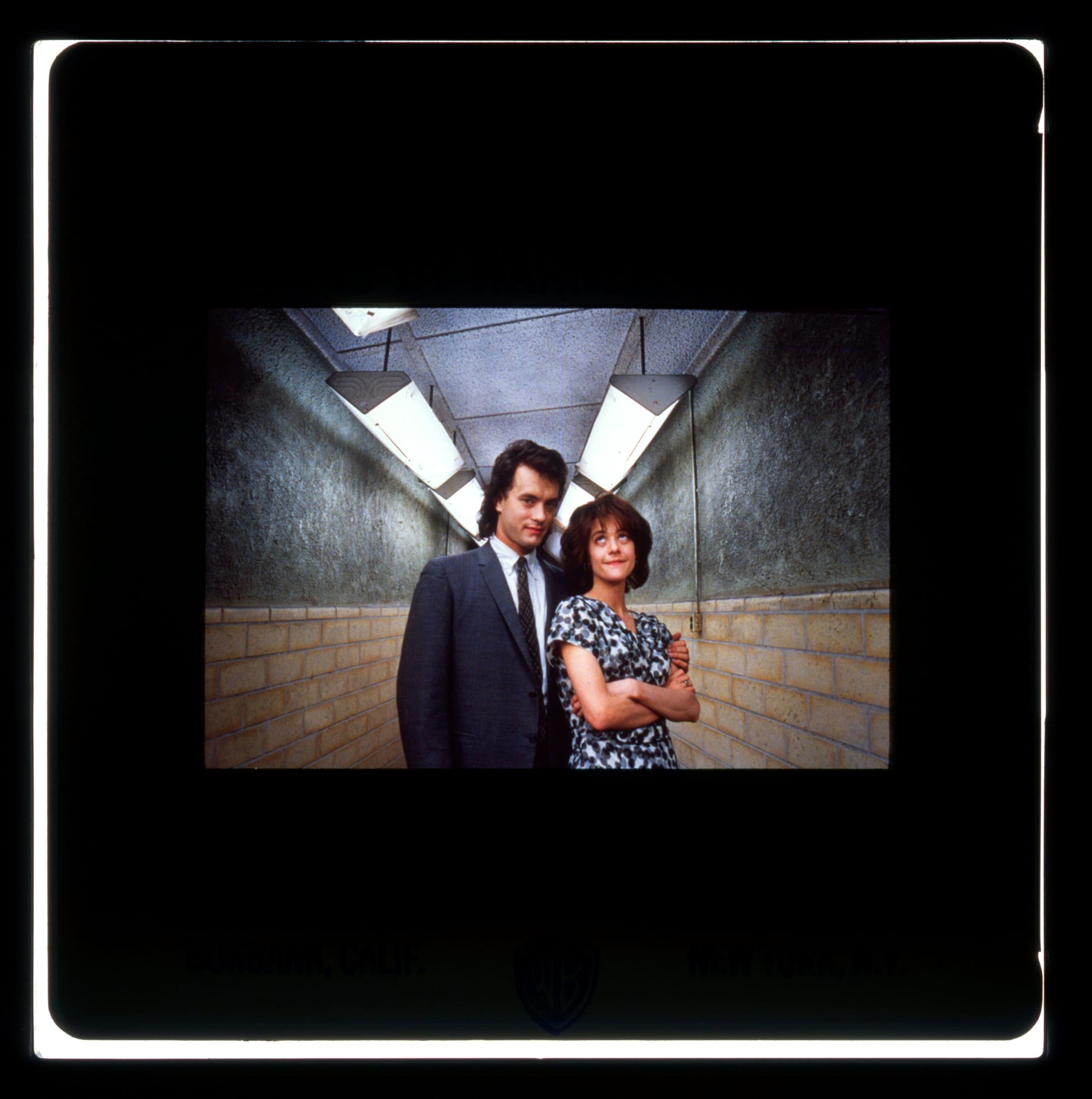 Color Publicity Slide: A man in suit and tie stands with arm around a woman in dress with arms folded and eyes looking up. They stand in a fluorescent lit hallway.