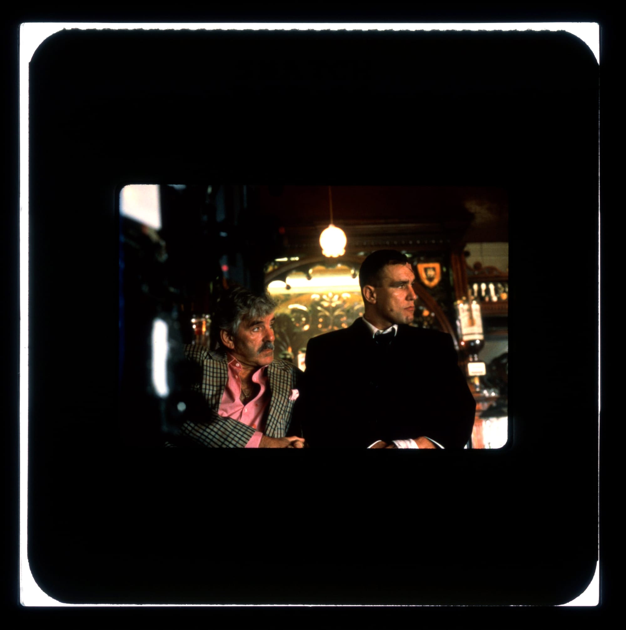 Color Publicity slide: Two men lean against a bar and look off-screen right with stern expressions.
