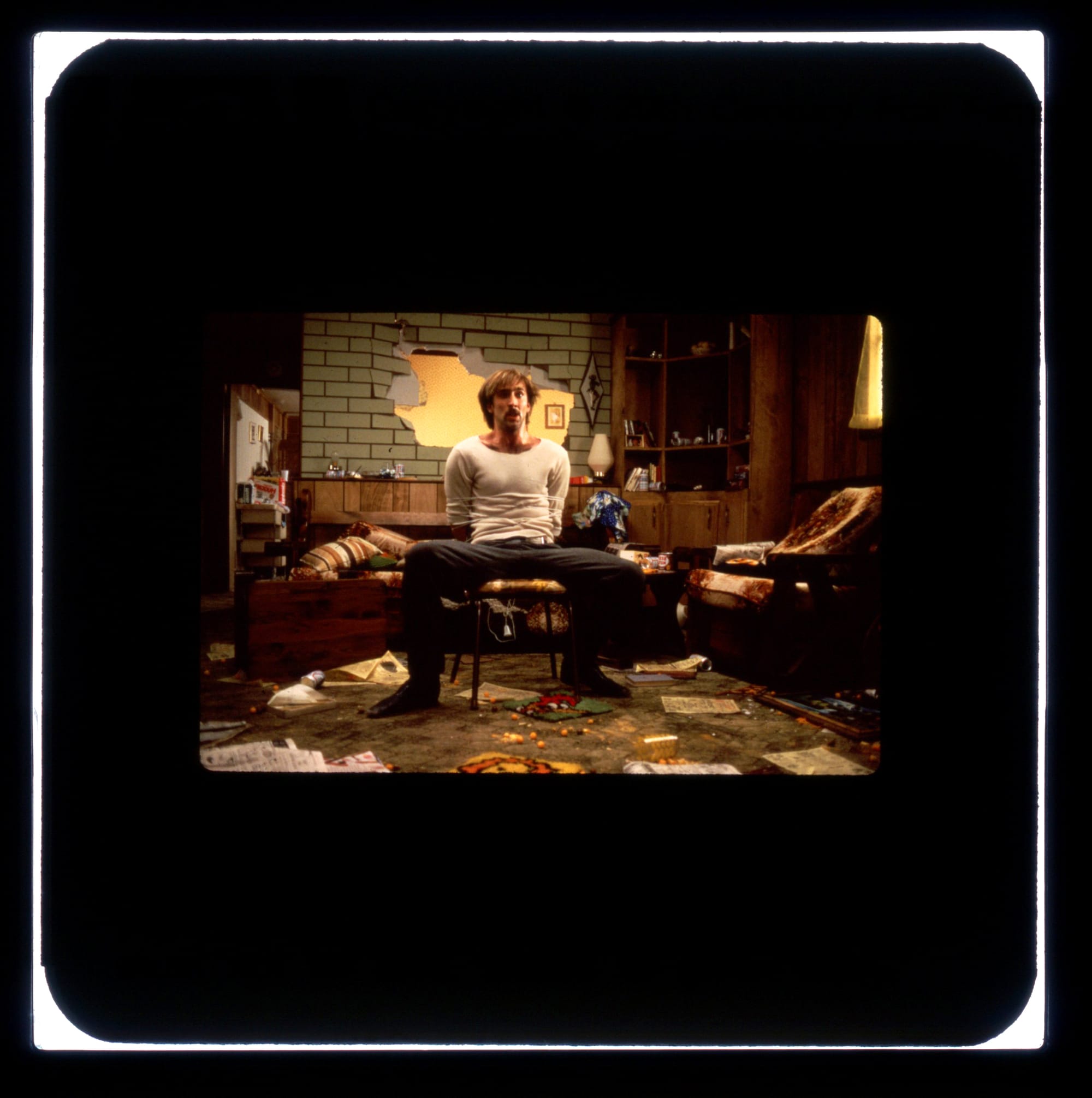 A man in a white shirt is tied to a chair with legs spread open. The room he's in is trashed with a large hole in the wall behind him.