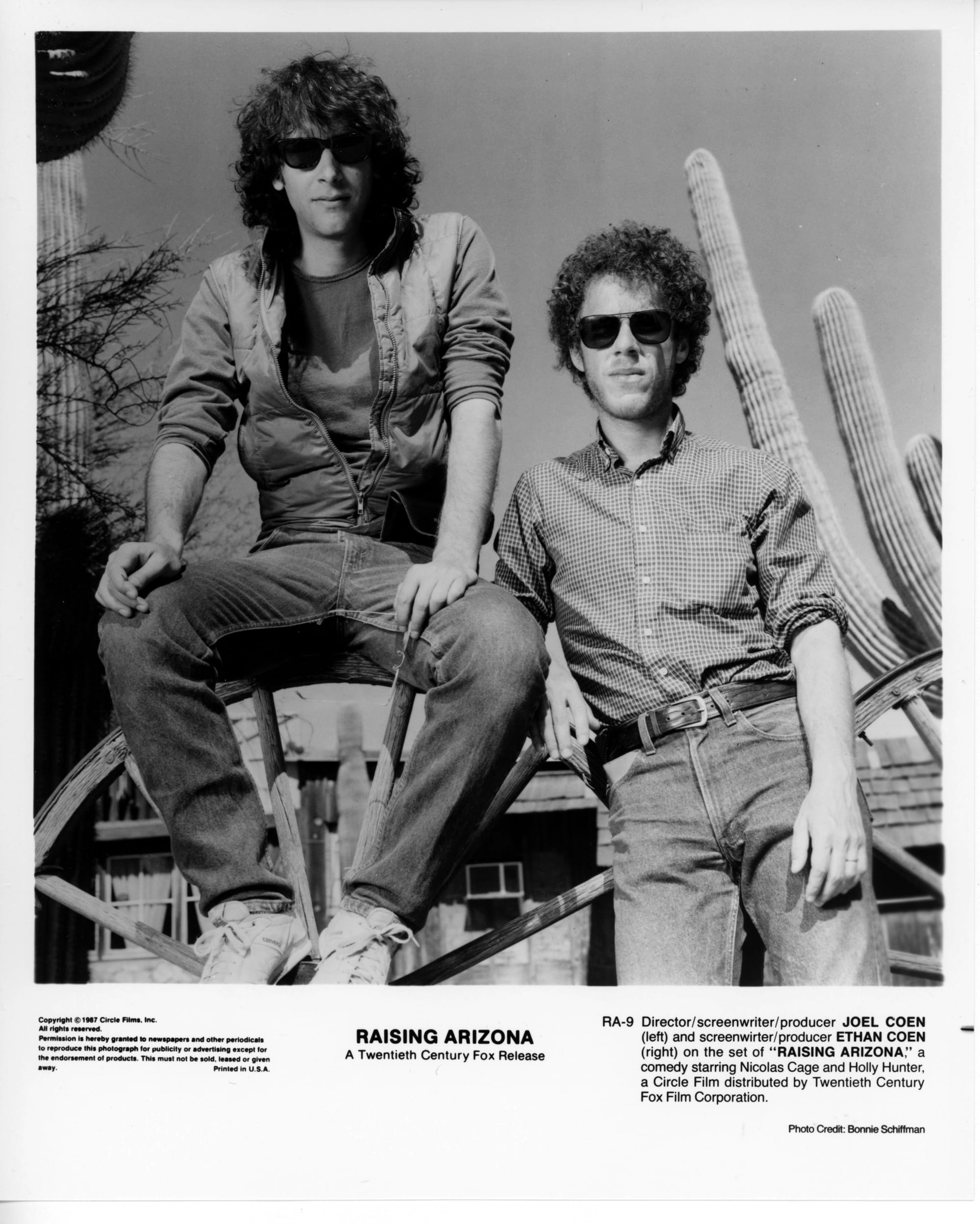 The Coen Brothers pose side by side outside with cacti behind them. Joel is on the left sitting atop a large wagon wheel. Ethan is on the right leaning against the same wheel. Both are wearing sunglasses and not smiling.