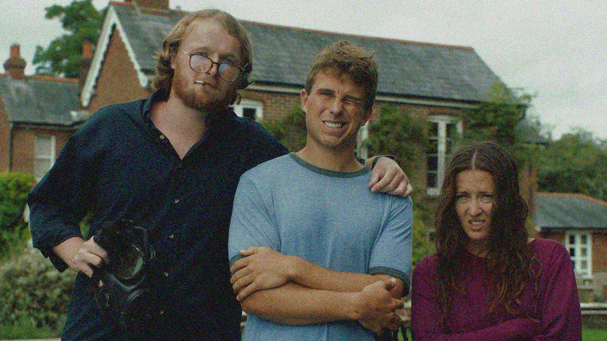 A man in glasses with a cigarette in mouth and gas mask in-hand poses with another man and woman for a photo. The latter two have more grimaces than smiles.
